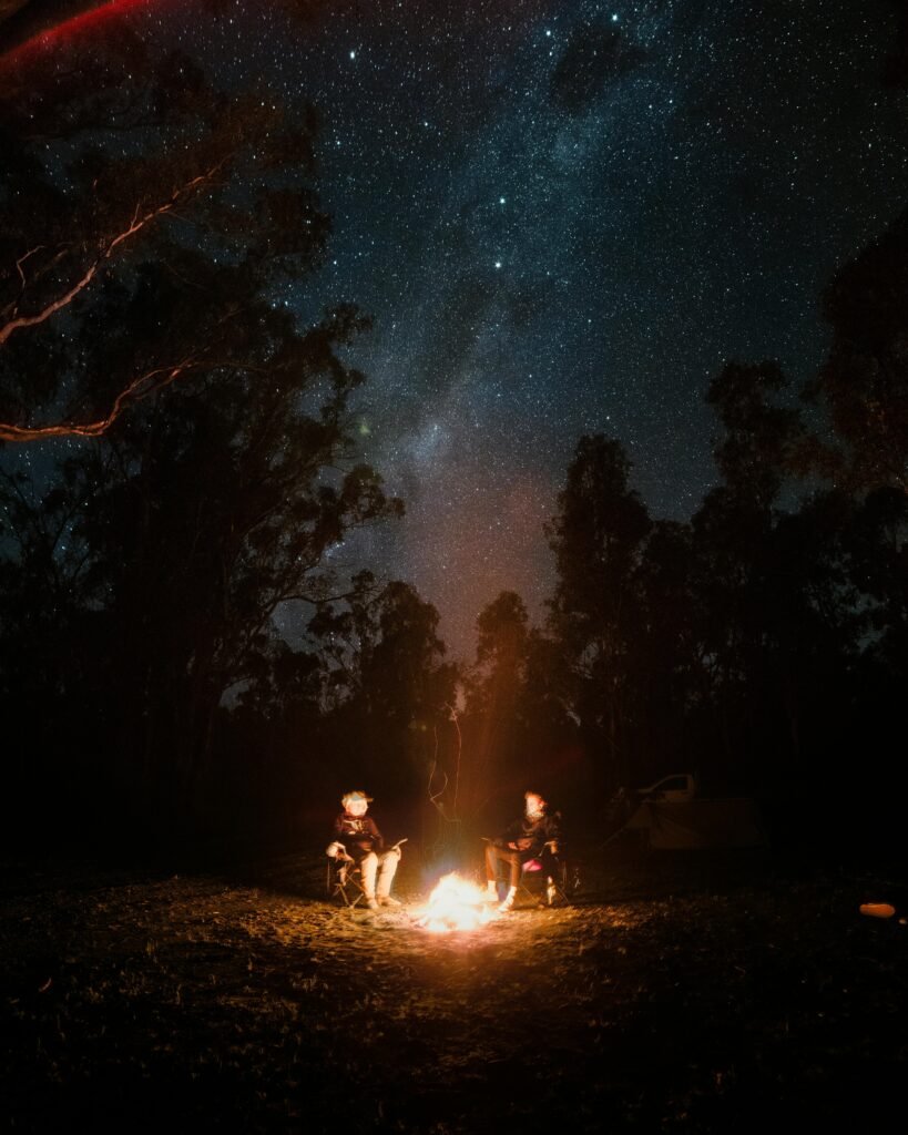 Two people enjoy stargazing by a campfire under a clear starry night sky, surrounded by trees.